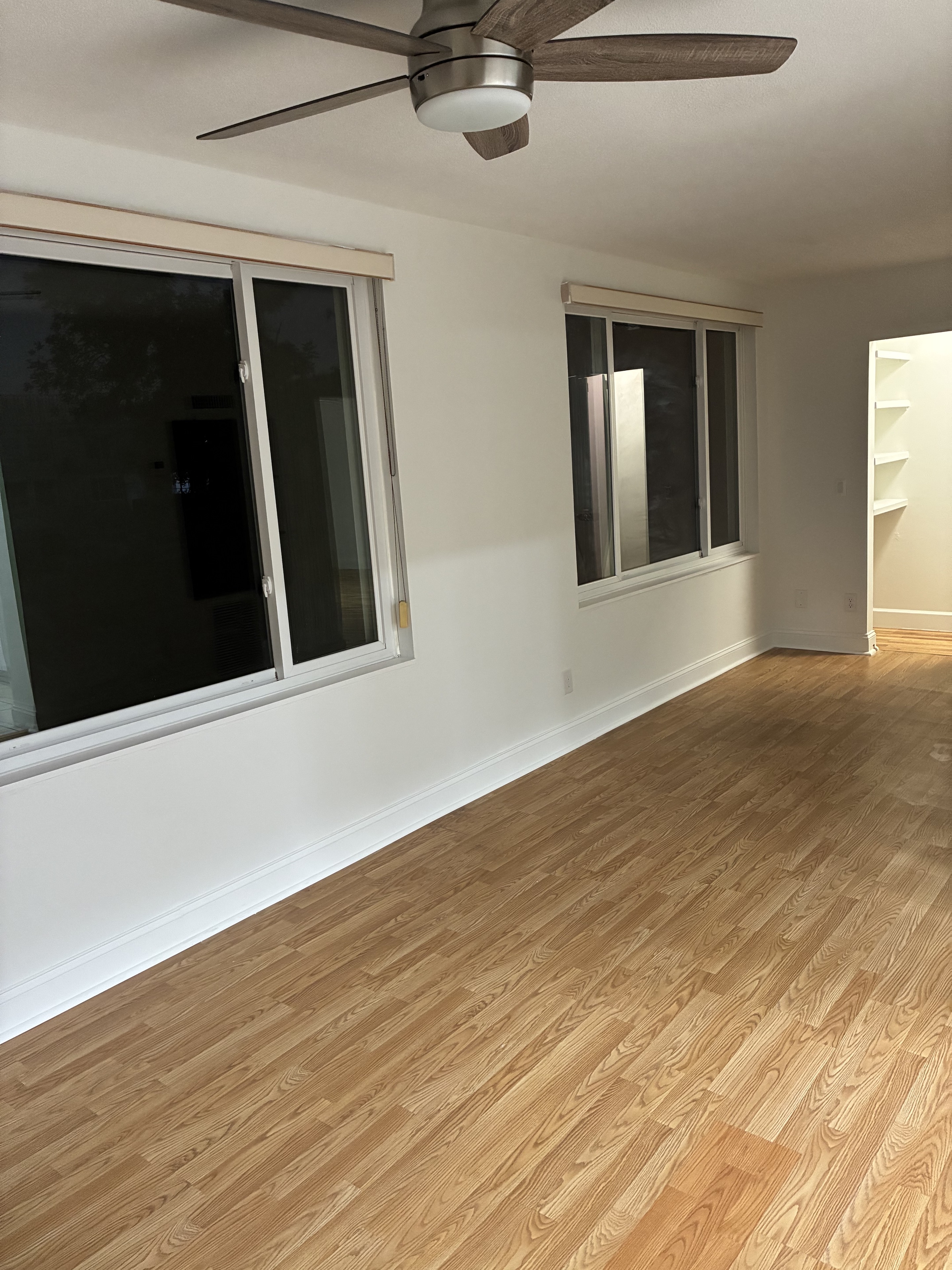 Clean empty bedroom with light hardwood floor, windows, and ceiling fan